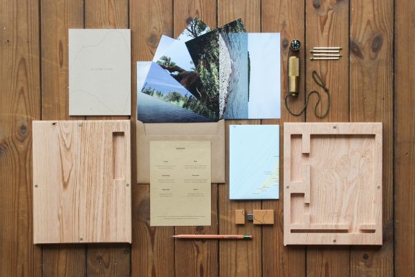 Publication laid out neatly on wood background featuring postcards, exhibition catalog, pencil, USB stick, and folded map between two pieces of CNC-machined red oak.
