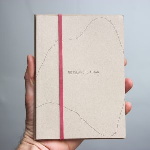 Hand holding a exhibition publication with the title No Island is Man in the center, surrounded by a thin line illustrating the precise coastline of an island. Cover is held closed with a vertical rubber band.